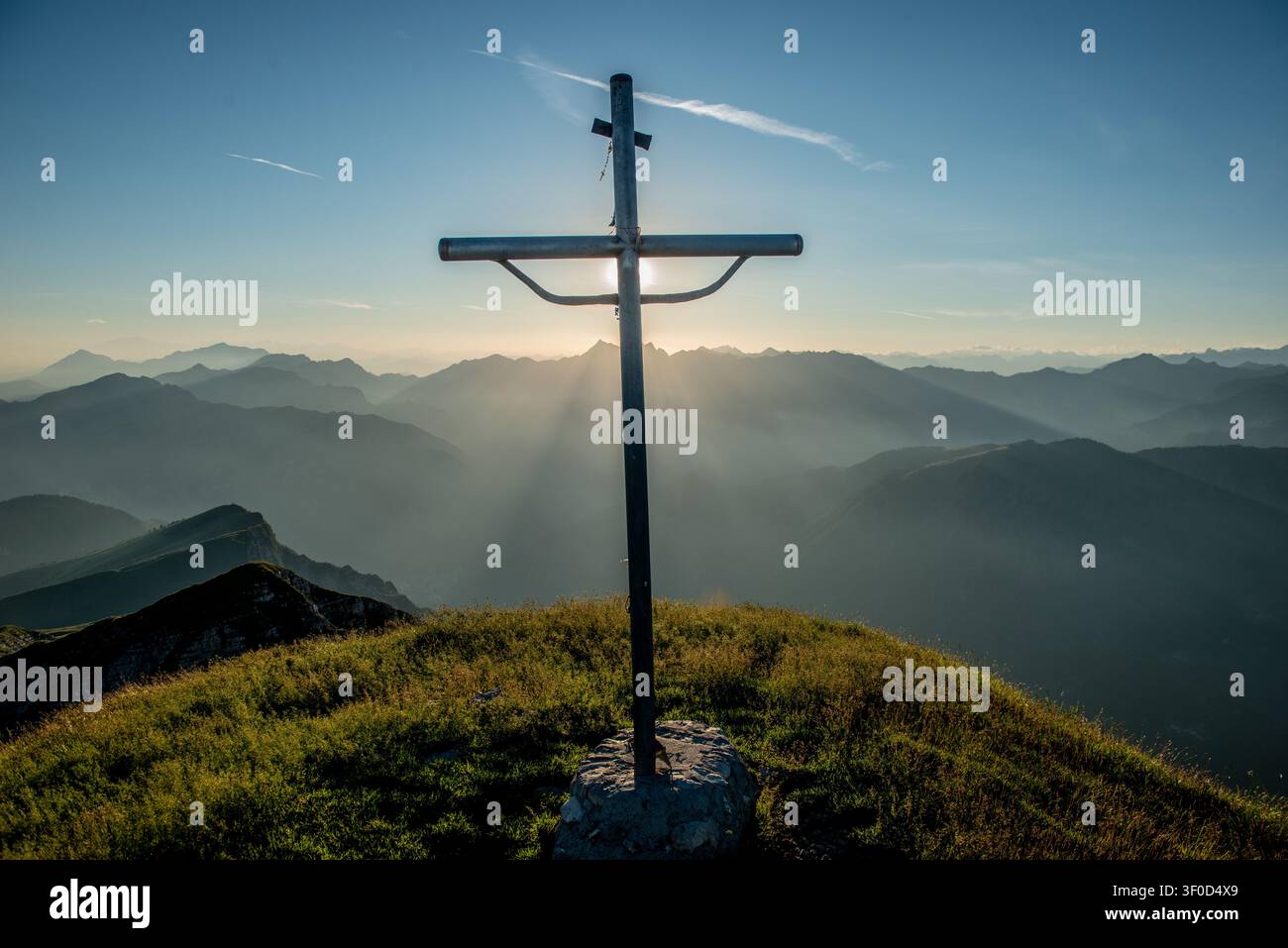 Die Strahlen der untergehenden Sonne dringen hinter dem Kreuz hervor und erzeugen einen Effekt göttlichen Lichts und von Spiritualität, der die umliegenden Gipfel umhüllt Stockfoto
