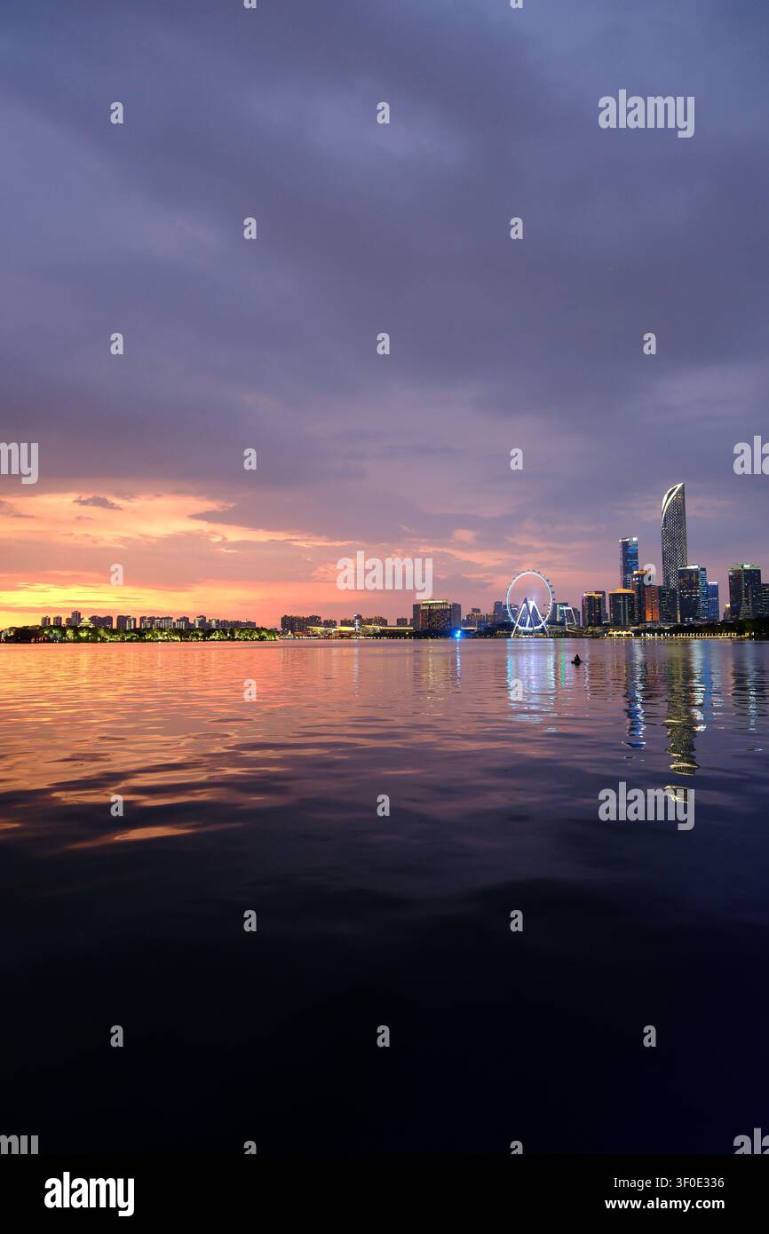 Ruhige Uferstadt-Skyline bei Sonnenuntergang mit lila Wolken, leuchtenden Lichtern, Reflexionen auf stillem Wasser und einem Riesenrad in der Ferne. Stockfoto