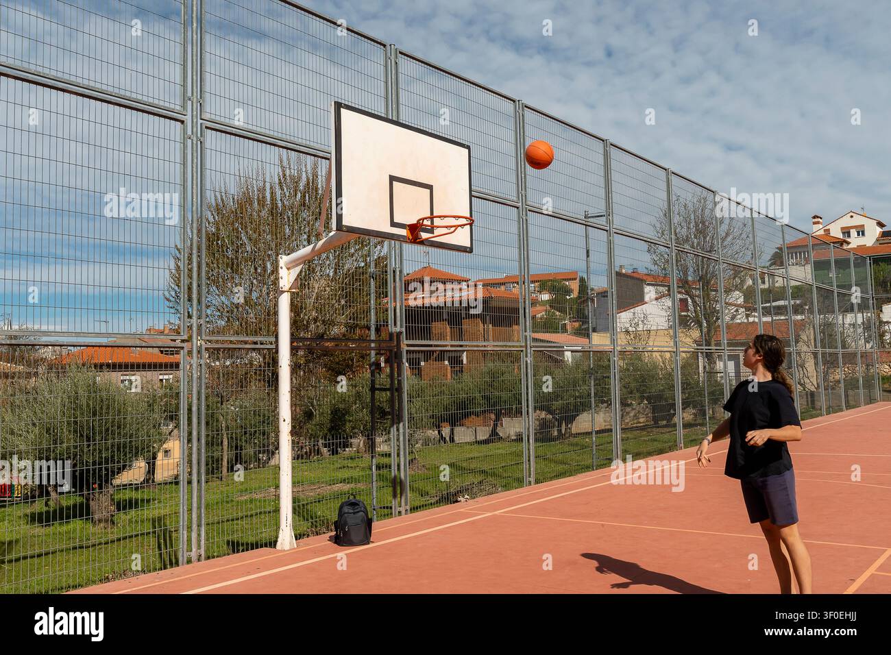 Ein Jugendlicher zielt auf den Korb, während er an einem sonnigen Tag auf einem Sportplatz mit Zäunen und Grünflächen Basketball spielt. Stockfoto