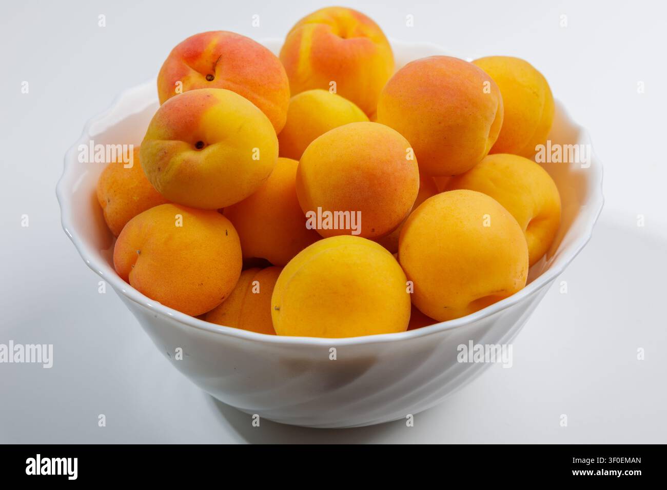 Es gibt ein Dutzend frische Aprikosen in einer tiefen weißen Schale. Die Früchte haben unterschiedliche Schattierungen – von hellgelb bis sattem Orange. Die Haut ist glänzend, slig Stockfoto