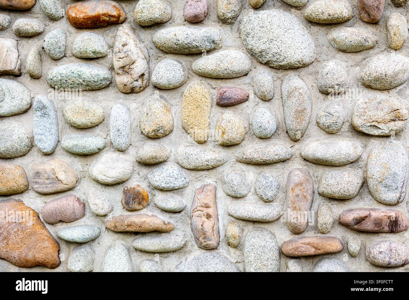 Abgerundete Kieselstein-Mauertextur. Abgerundete Kieselsteine, die in Mörtel gesetzt sind, bilden eine strukturierte Steinmauer mit grauen, beigen und braunen Natursteinmustern. Stockfoto