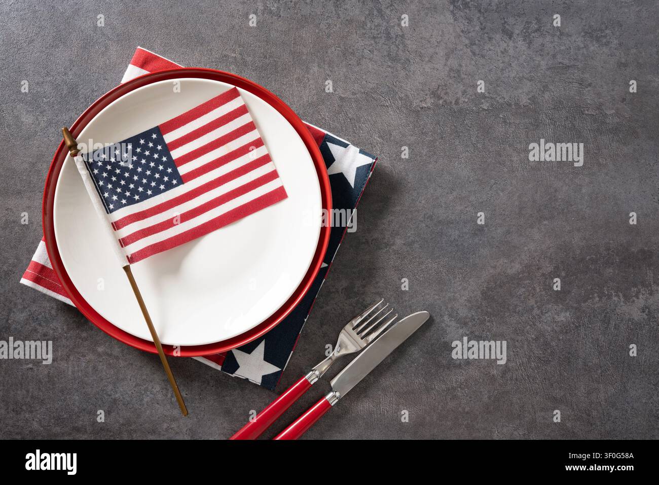 Weißer Essteller mit amerikanischer Flagge patriotischem Serviette und rotem Besteck auf grauem Hintergrund mit Kopierraum. Ansicht von oben. Stockfoto