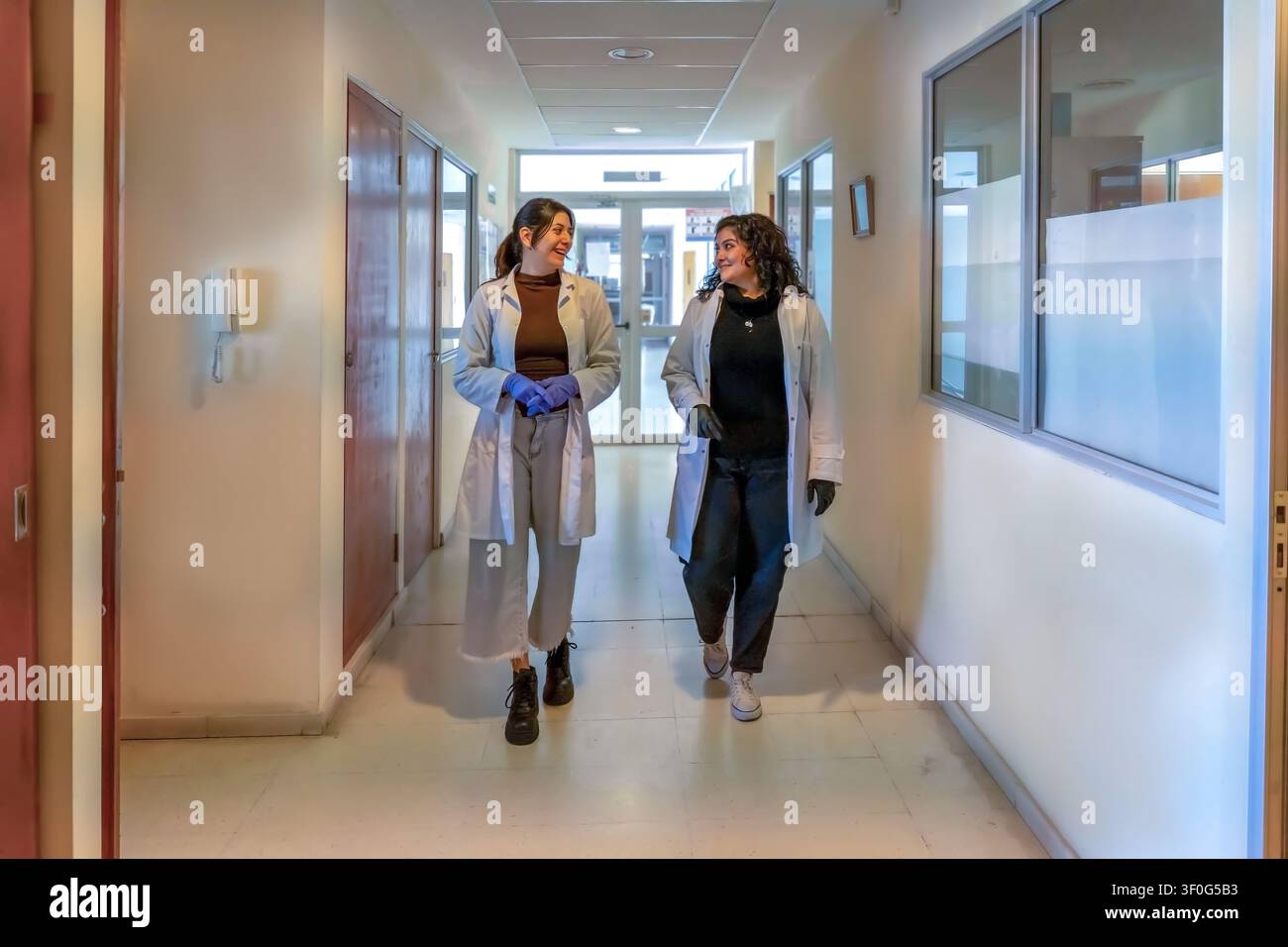 Zwei junge Wissenschaftlerinnen in weißen Laborkitteln und Handschuhen, die lächeln und sich unterhalten, während sie zusammen durch einen hellen institutionellen Korridor mit Tür gehen Stockfoto