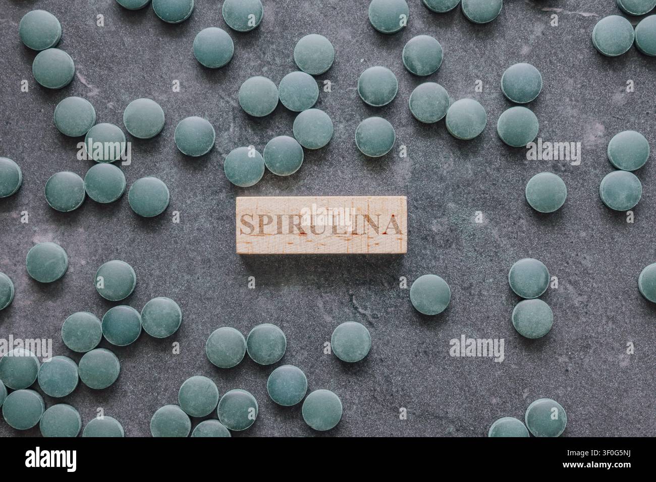 Horizontale Aufsicht-Aufnahme von grünen Spirulina-Chlorella-Algen-Tabletten, die auf einem dunkelgrauen Steinboden um ein kleines hölzernes Schildblock verstreut sind Stockfoto
