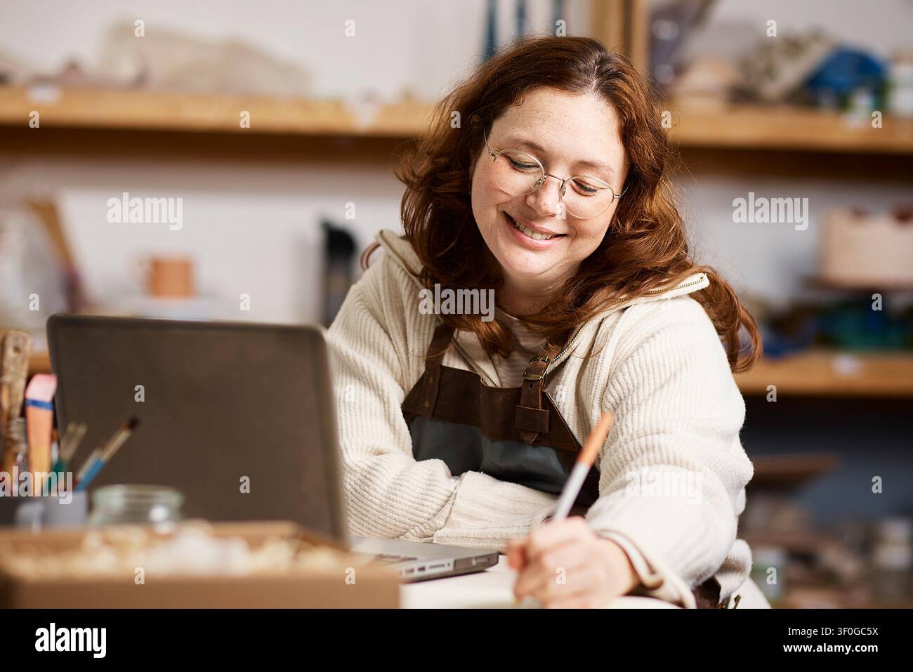 Lächelnde Töpferin arbeitet in ihrem Töpferatelier mit Laptop und Werkzeugen, während sie ein kleines Unternehmen führt Stockfoto