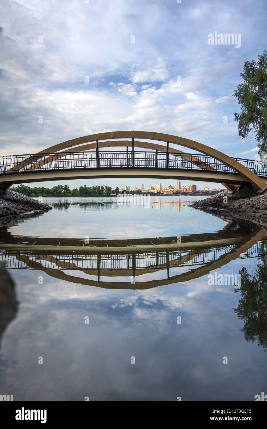 Fußgängerbrücke in Vilnius im Obolon-Bezirk von Kiew, Ukraine, die sich an einem friedlichen Sommermorgen im ruhigen Wasser spiegelt Stockfoto