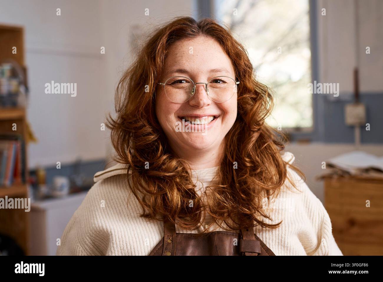 Lächelnde rothaarige Töpferwerkstatt-Besitzerin mit Schürze und Brille in kreativem Kleinunternehmens-Workshop Stockfoto