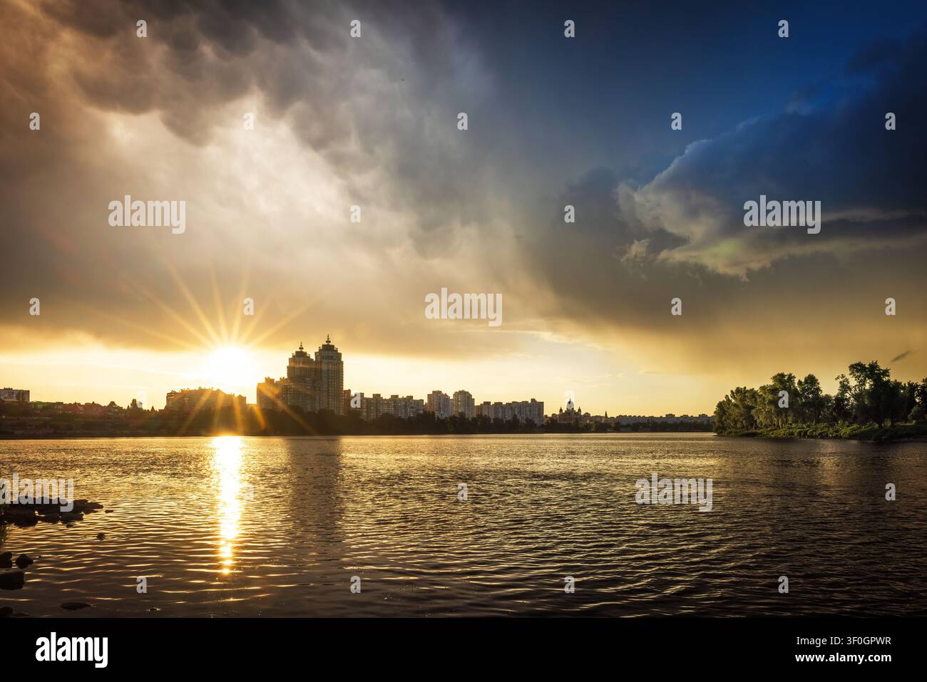 Dramatischer Sonnenuntergang nach einem Gewitter über der Obolon-Uferpromenade in Kiew, Ukraine. Goldene Sonnenstrahlen brechen durch die Gewitterwolken und spiegeln sich im Dnipro wider Stockfoto