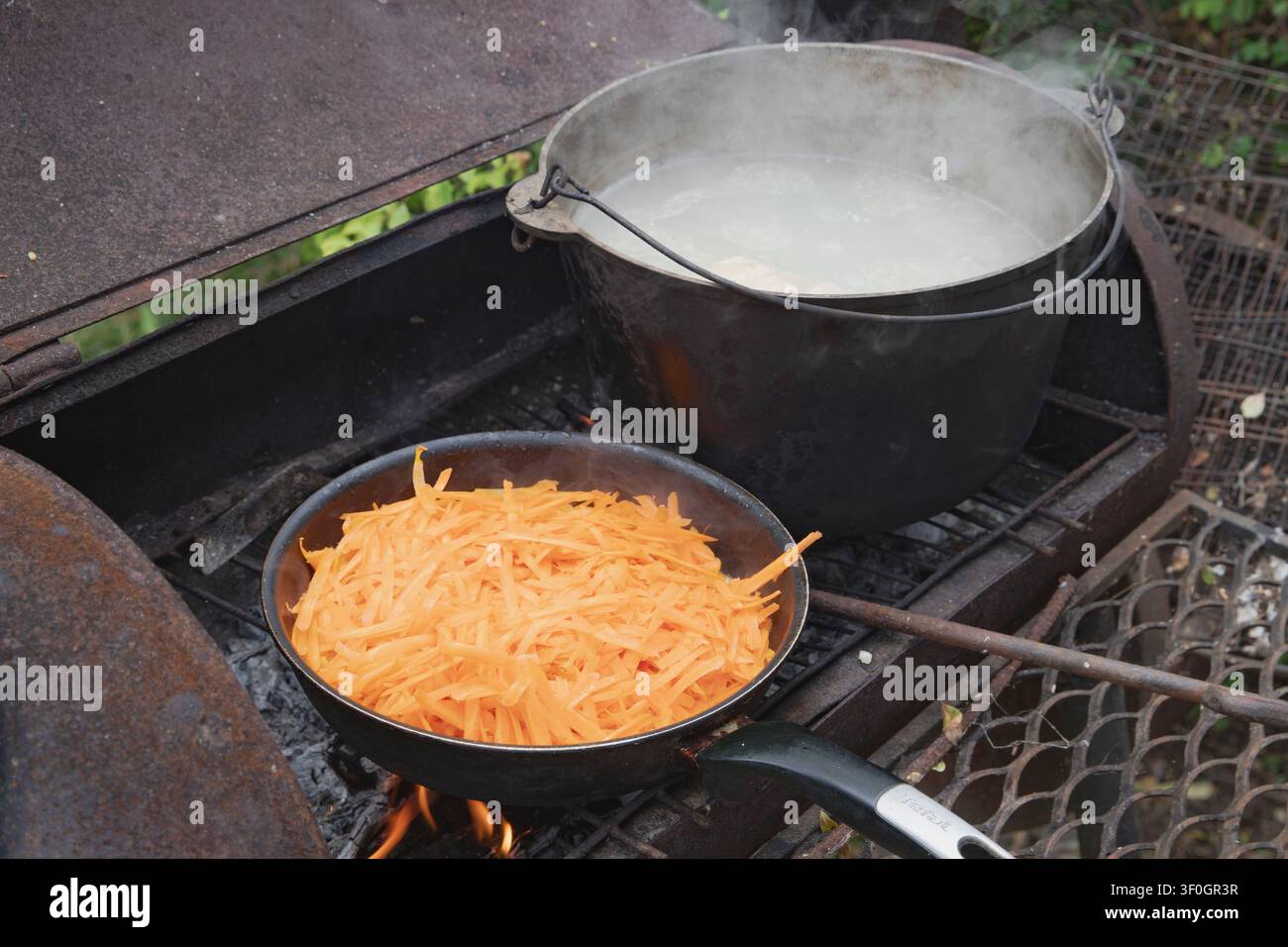 Geriebene Karotten braten in der Pfanne neben einem kochenden Topf auf einem rustikalen Holzofen im Freien Stockfoto