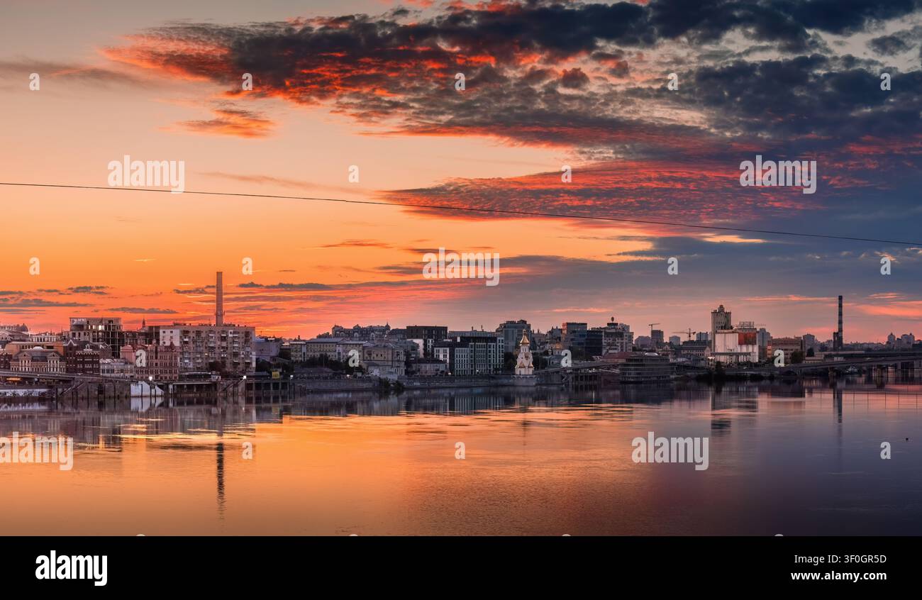 Nachtansicht des Podil-Bezirks in Kiew, Ukraine, mit dem Dnipro-Fluss, der Uferpromenade und einem farbenfrohen Sonnenuntergang, der im Abendhimmel verblasst. Stockfoto