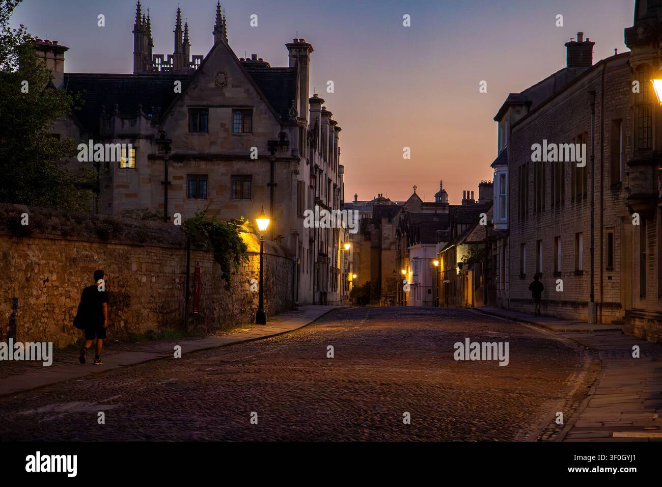 Eine einsame Gestalt geht am Abend entlang der gepflasterten Merton Street in Oxford, während warme Straßenlaternen die historischen Kolleggebäude beleuchten. Stockfoto