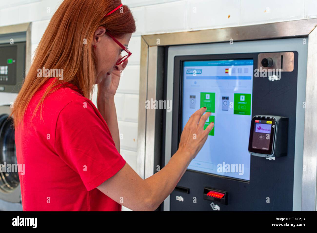 Frau mit roten Haaren und Brille, die Waschmaschinenoptionen auf einem digitalen Touchscreen in einem modernen Selbstbedienungswaschkeller auswählt, Haushaltsdinge verwaltet Stockfoto