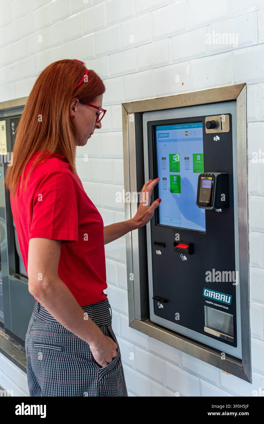 Frau berührt einen modernen Selbstbedienungs-Zahlungs- und Auswahl-Touchscreen-Automaten in einer Wäscherei und wählt Optionen für automatisches Waschen und Trocknen aus Stockfoto
