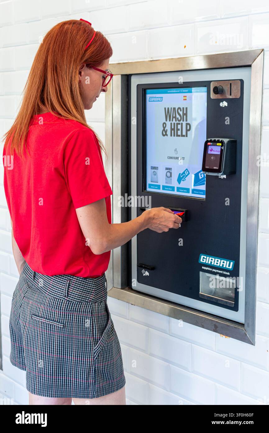 Rotehaarige Frau, die eine Kreditkarte in ein Zahlungsterminal einführt, bedient einen Selbstbedienungs-Wasch- und Trocknerkiosk für saubere weiche Kleidung in einer Mo Stockfoto