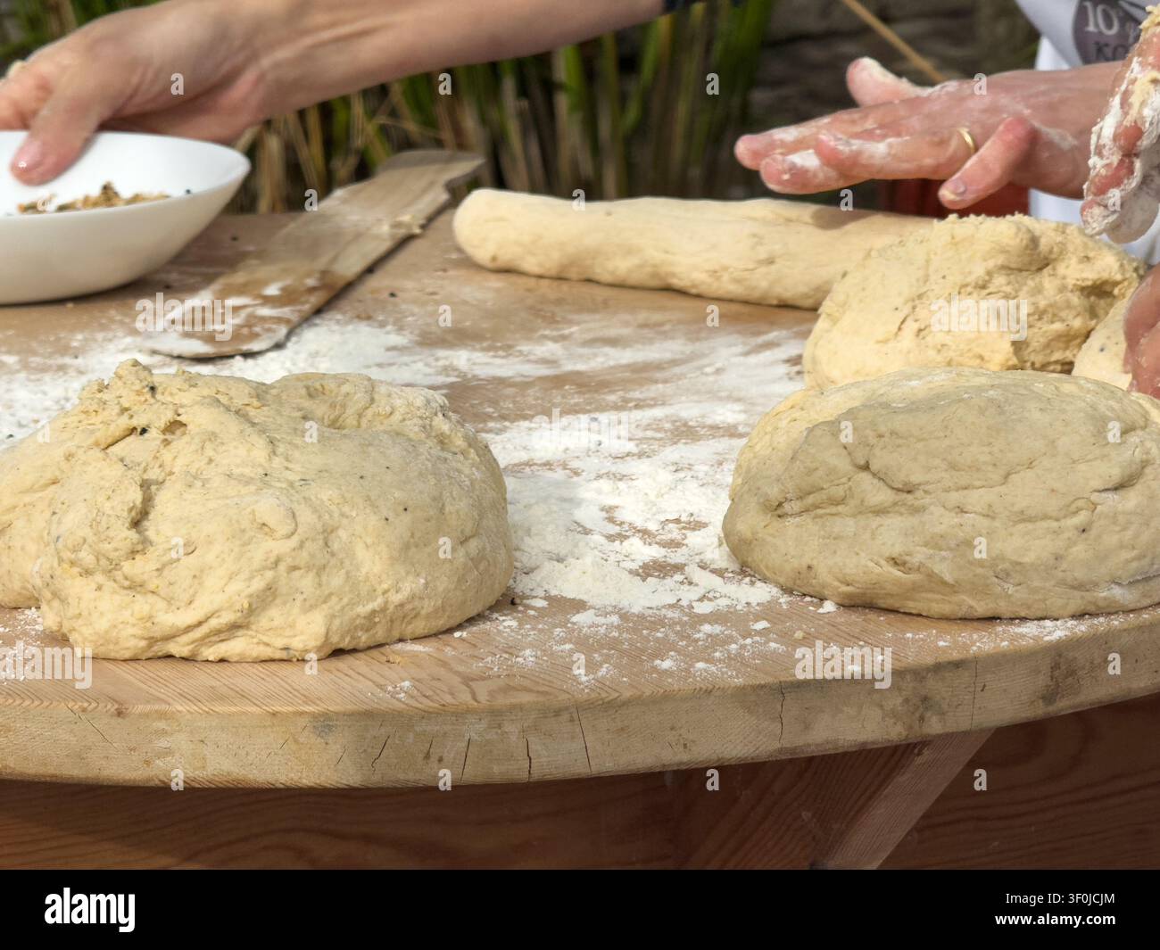 Hände, die traditionellen kretischen Brotteig auf einem rustikalen Holztisch zubereiten, der mit Mehl bestreut ist, während eines authentischen Backworkshops im Freien. Stockfoto