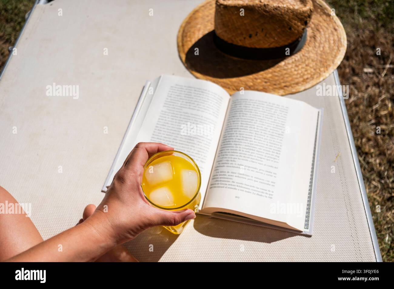 Eine erfrischende Szene sommerlicher Entspannung mit einem offenen Buch und einem kalten Getränk mit Eiswürfeln an einem sonnigen Tag. Ein Strohhut liegt daneben und rundet die Szene ab Stockfoto