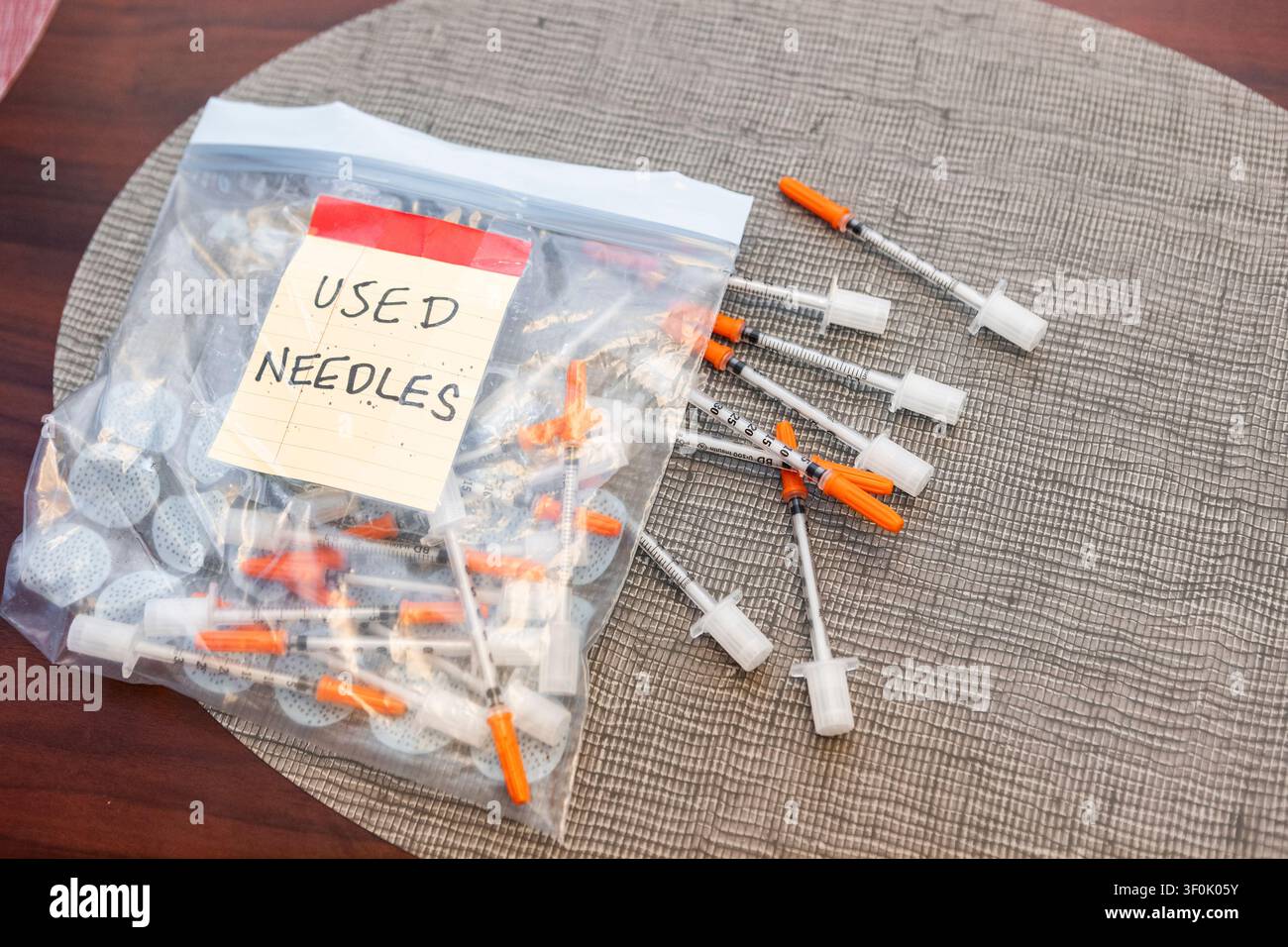 Ein Ziplock-Beutel gefüllt mit gebrauchten Insulin-Spritzen und -Nadeln, neben anderen, die auf einer strukturierten Matte liegen. Diese Anordnung betont richtige Entsorgungsmethoden Stockfoto