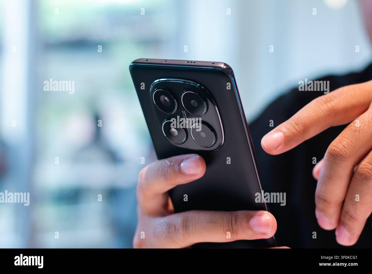 Eine Nahaufnahme, die eine menschliche Hand zeigt, die ein schlankes schwarzes Mobilgerät mit einem markanten Dreifach-Kamera-Setup auf der Rückseite hält, drinnen bei weichem Licht verwendet Stockfoto