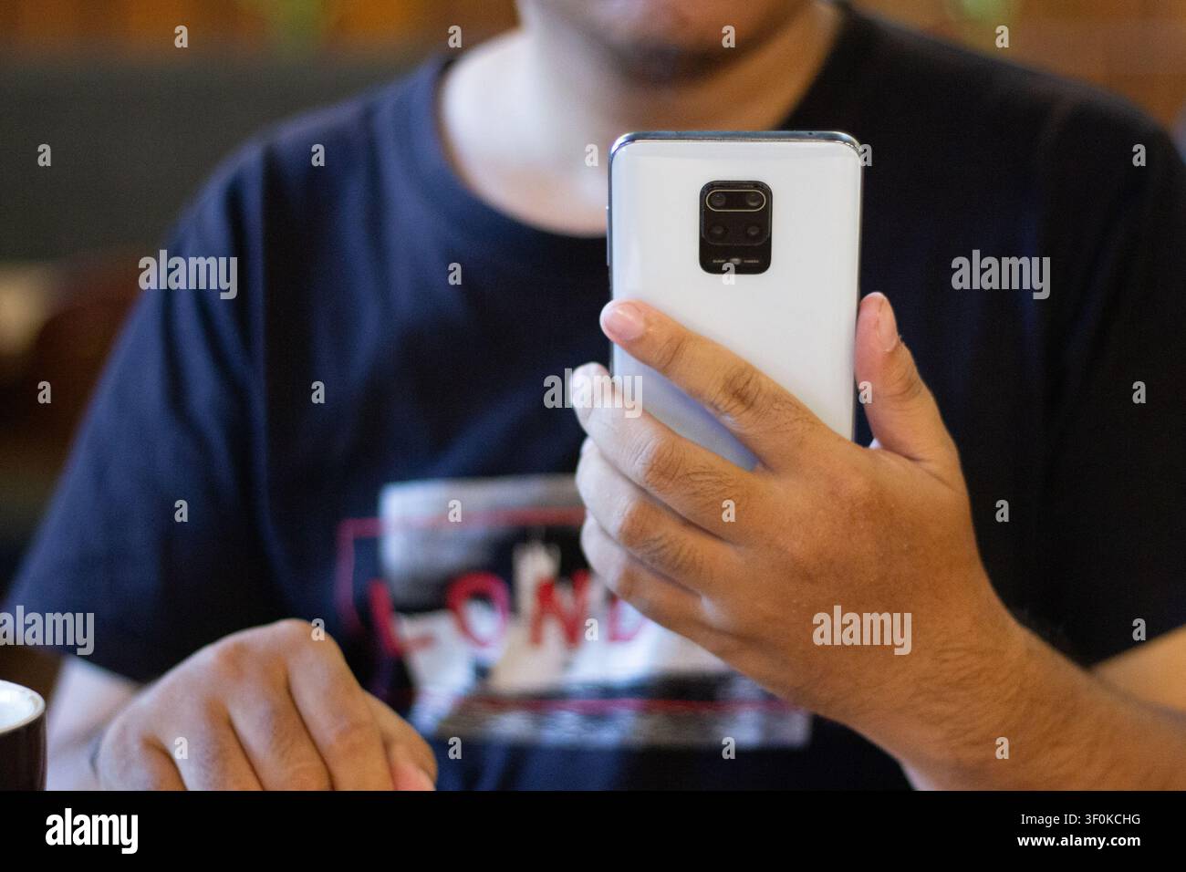 Eine Nahaufnahme zeigt eine lässig gekleidete Person, die ein Mobilgerät in einer lebendigen Kaffeeschankatmosphäre hält, wobei weiches Licht die urb Stockfoto