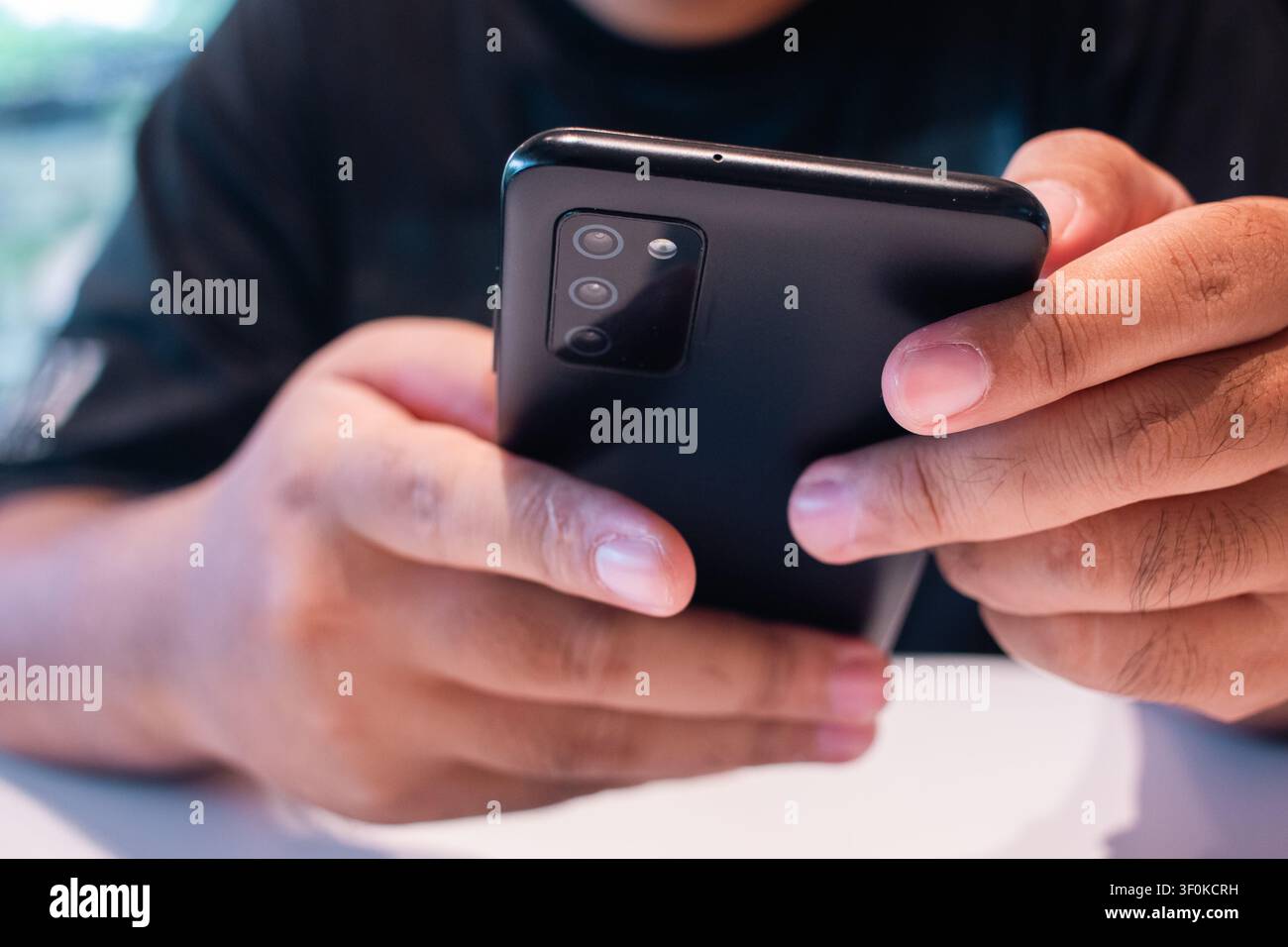 Eine konzentrierte Person hält ein Mobilgerät in den Händen, eingebettet in eine sanft beleuchtete Innenumgebung, die die Geste der Interaktion betont Stockfoto