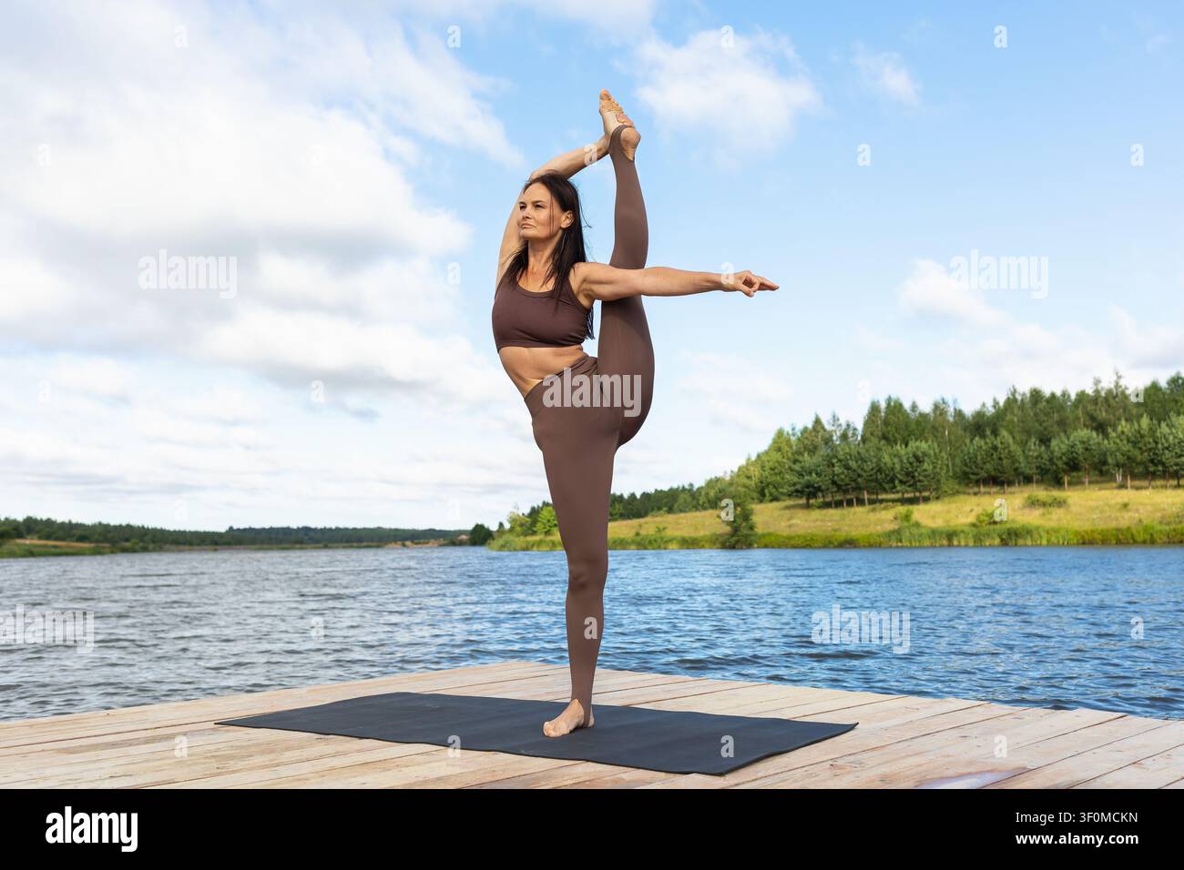 Frau, die im Stehen den vertikalen Spagat-Yoga-Posen Urdhva Prasarita Eka Padasana auf einem Steg am See an einem sonnigen Sommermorgen ausführt Stockfoto