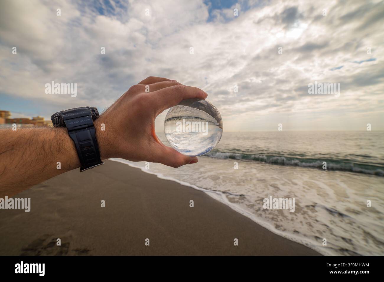 Eine Hand, die eine taktische Solaruhr trägt, hält eine klare Kristall-Linsen Kugel auf Augenhöhe an einem sandigen Meeresstrand. Stockfoto