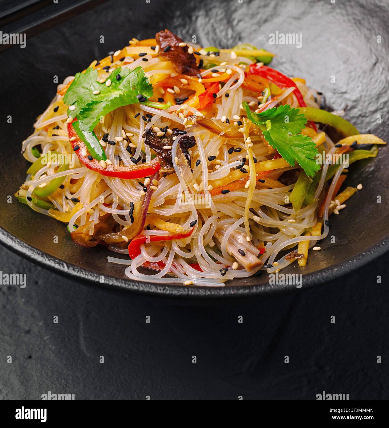 Glasnudeln-Stir-Fry mit bunten Gemüsen, Pilzen und Sesamsamen in einer schwarzen Schüssel Stockfoto