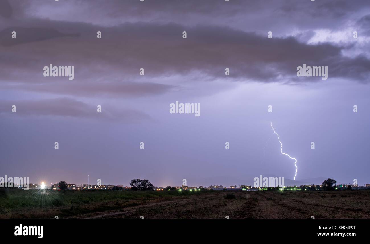 Ein mächtiger Blitzschlag vom Wolkenboden trifft in der Nähe des Randes einer mittelgroßen Stadt, aufgenommen von einer offenen landwirtschaftlichen Ebene in der Dämmerung. Stockfoto
