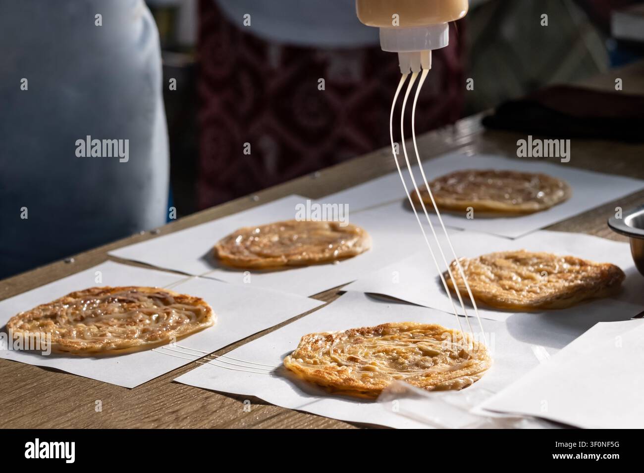 Hand, die Kondensmilch über knusprige Roti-Fladenbrote gießt, die auf Papier mit einer Quetschflasche liegen, Nahaufnahme Stockfoto
