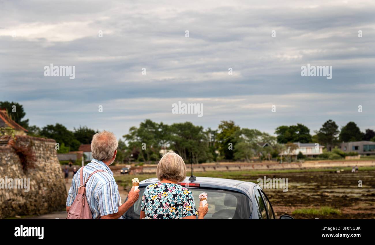 Ein älteres Paar von hinten gesehen, genießt Eiscremewaffeln auf der Shore Road, während es über die bei Ebbe trocken liegenden Schlickflächen des Bosham Creek blickt. Stockfoto