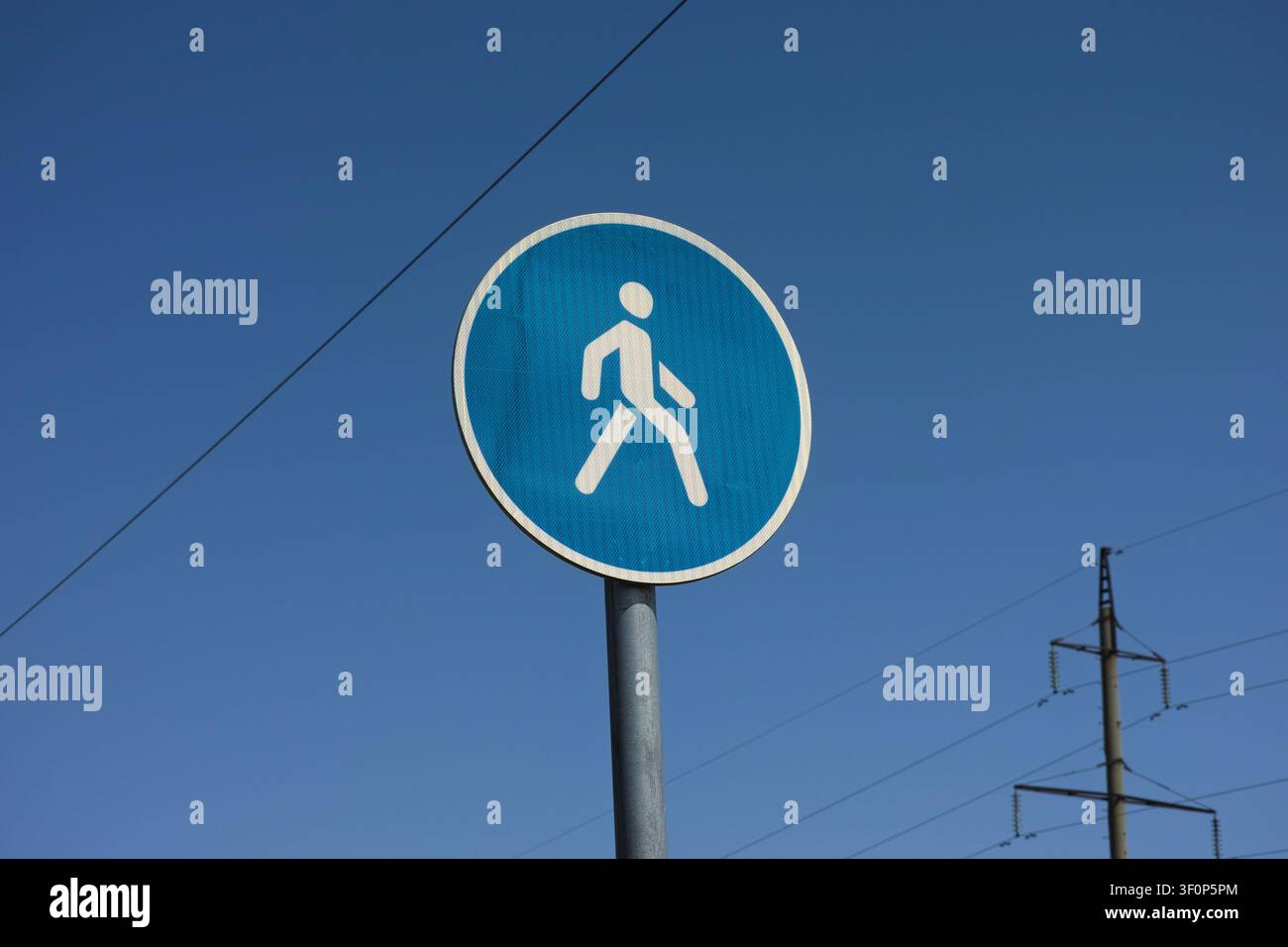 Ein rundes blaues Fußgängerweg-Verkehrsschild auf einem Metallpfosten vor einem blauen Himmel mit Stromleitungen. Stockfoto