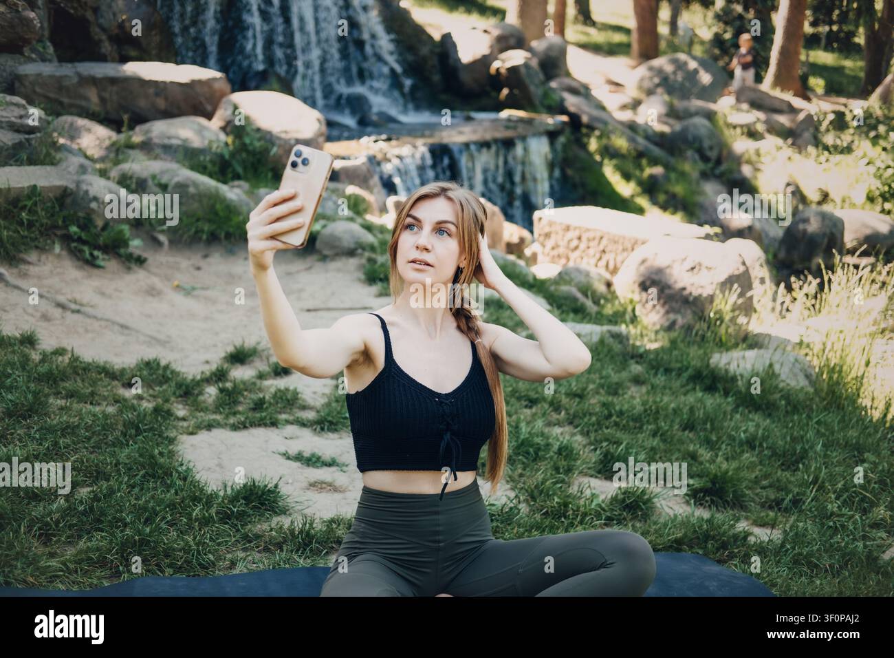 Junge erwachsene Frau sitzt auf Yogamatte und hält Smartphone, Wasserfall und Felsen hinter ihr. Digitales Wohlbefinden, Outdoor-Aktivität, Selbstfürsorge, Natur Stockfoto