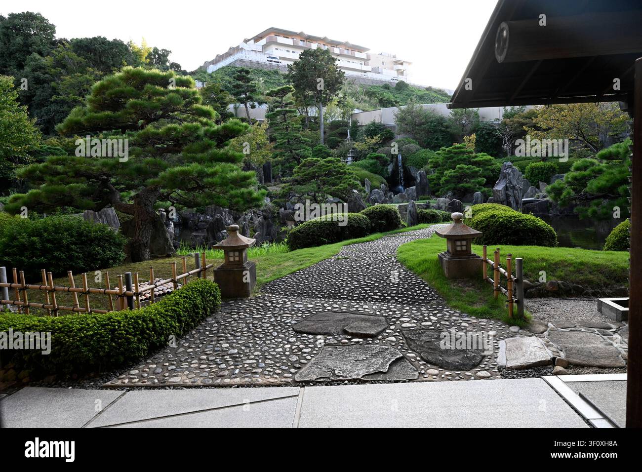 Steinpflasterweg, flankiert von traditionellen Steinlaternen, der durch den gepflegten Hanbe-Garten führt, einen bekannten japanischen Garten, der eine Luxusanlage umgibt Stockfoto