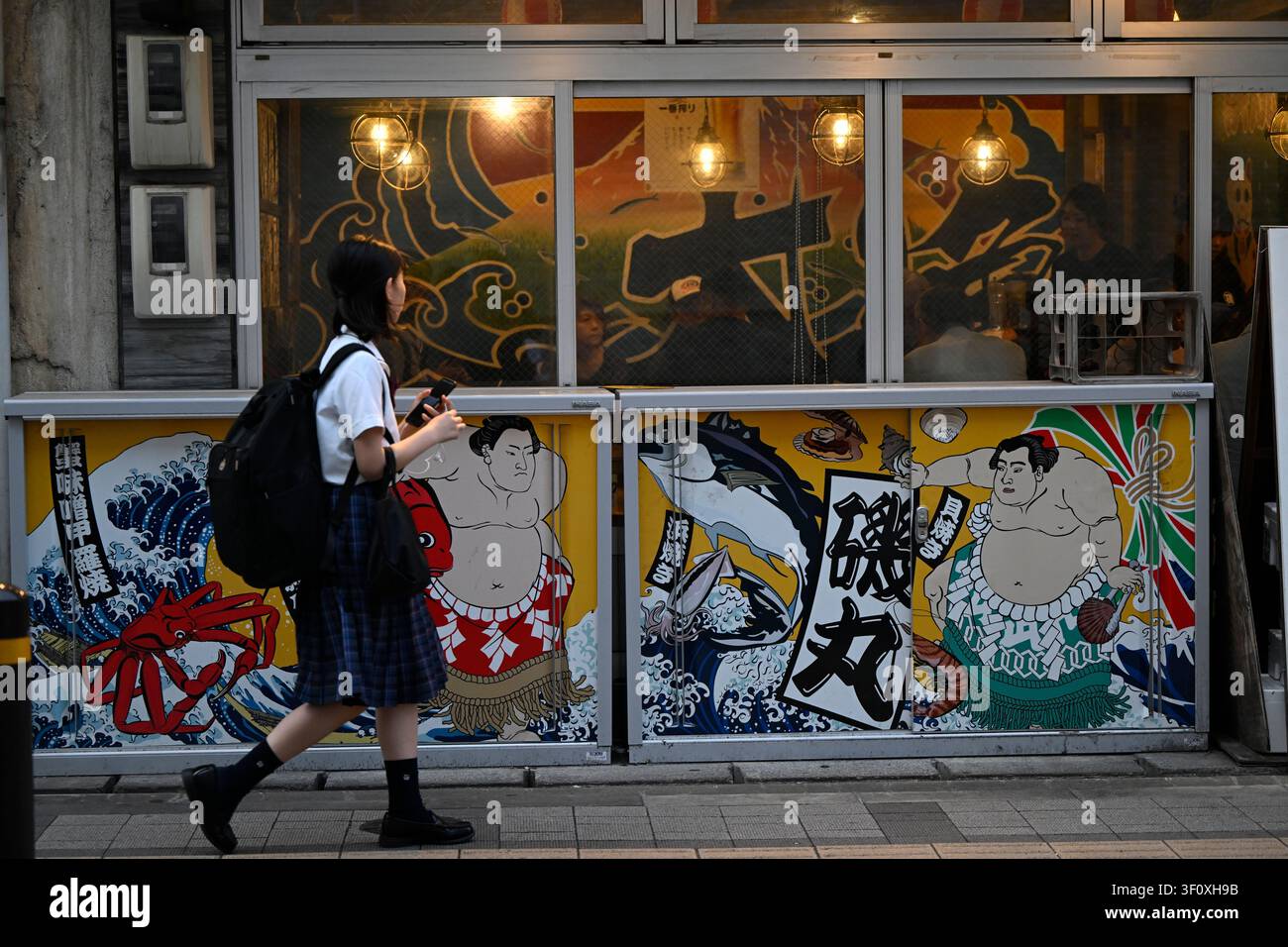 Tokio, Japan – Mai 2026: Eine Schulmädchen geht an einem Fischereirestaurant vorbei, das mit bunten Sumoringer-Illustrationen und traditioneller japanischer Gra Stockfoto