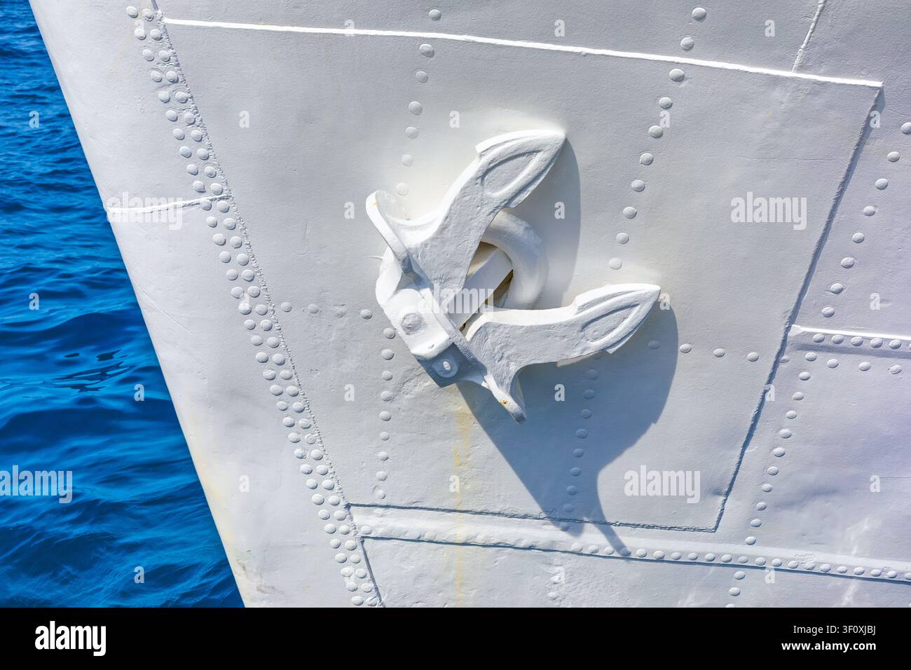 Weiß gestrichener Anker liegt gesichert gegen einen genieteten Schiffsboden, seine klare geometrische Form steht in scharfem Kontrast zum lebhaften blauen Meer darunter Stockfoto