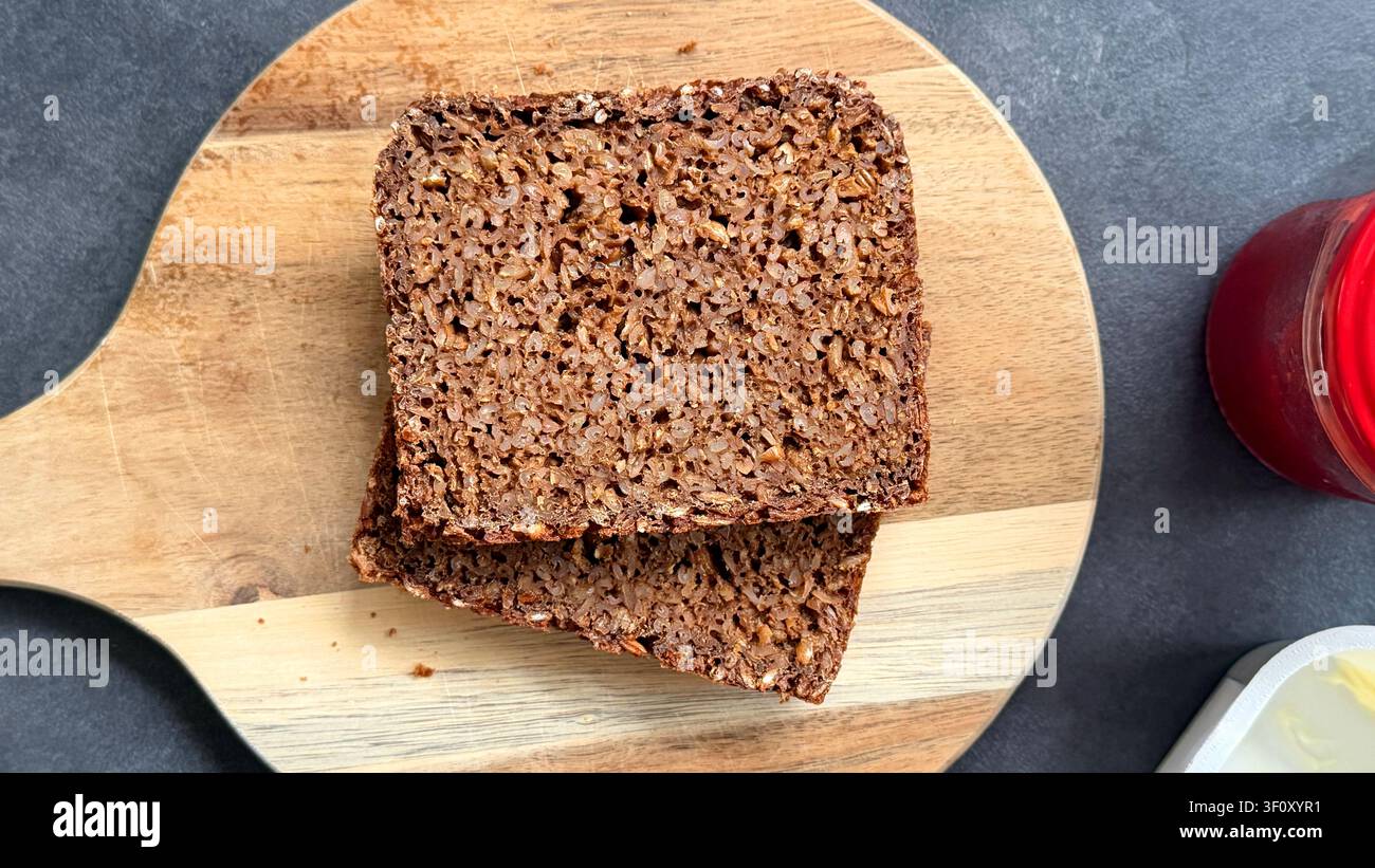 Scheiben dunkles Saatenbrot auf einem Holzschneidebrett mit einem Glas Marmelade und Butter auf einer grauen Arbeitsplatte Stockfoto