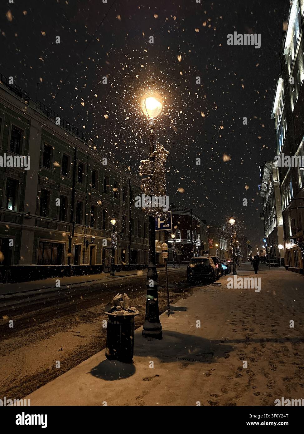 Schneefall, der von einer Straßenlaterne auf einer ruhigen historischen Straße bei Nacht beleuchtet wird - Smartphone-aufgenommenes Stockfoto