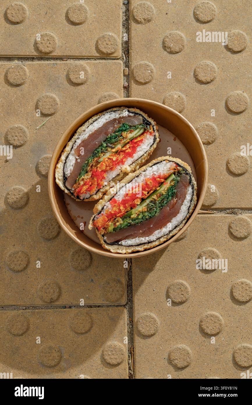 Sushi-Burger auf gelben texturierten Pflastersteinen. Lieferbox. Isoliert. Kunstvolles Foto. Professionelles Studiolicht. Stockfoto