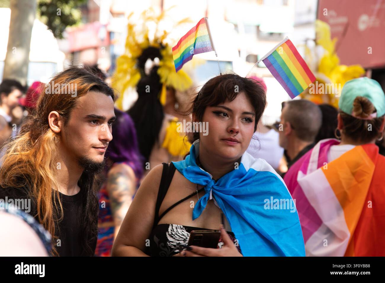 Zwei Personen stehen zusammen bei einer Pride-Parade, eine trägt kleine Regenbogenflaggen in ihren Haaren und einen hellblauen Schal, umgeben von einer farbenfrohen Menschenmenge Stockfoto