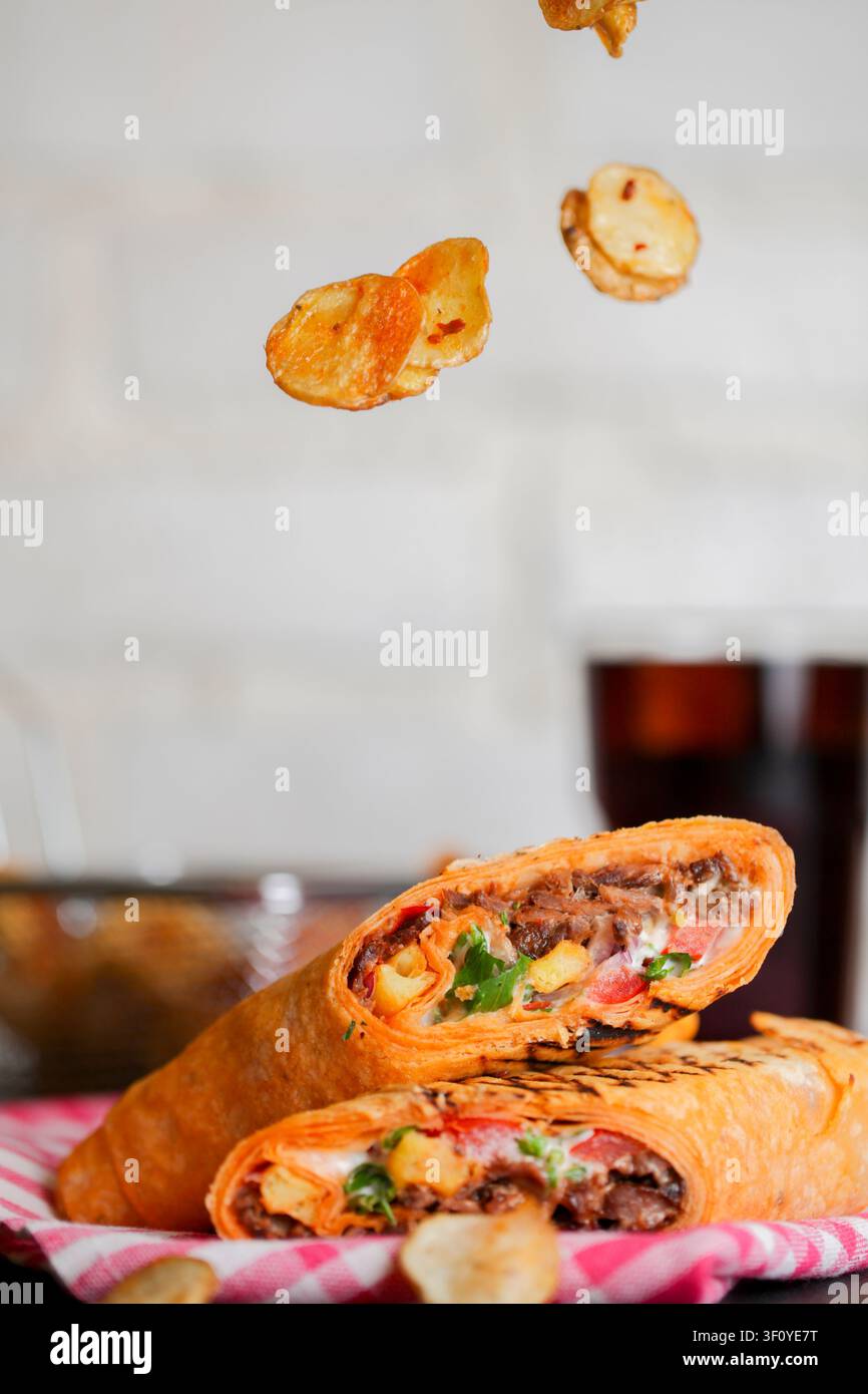 Zwei Wrap mit Fleisch und Gemüse stehen auf einem Tisch. Kartoffelchips fallen von oben. Ein Getränk steht in der Nähe. Die Szene zeigt die Zubereitung von Speisen und Genuss Stockfoto