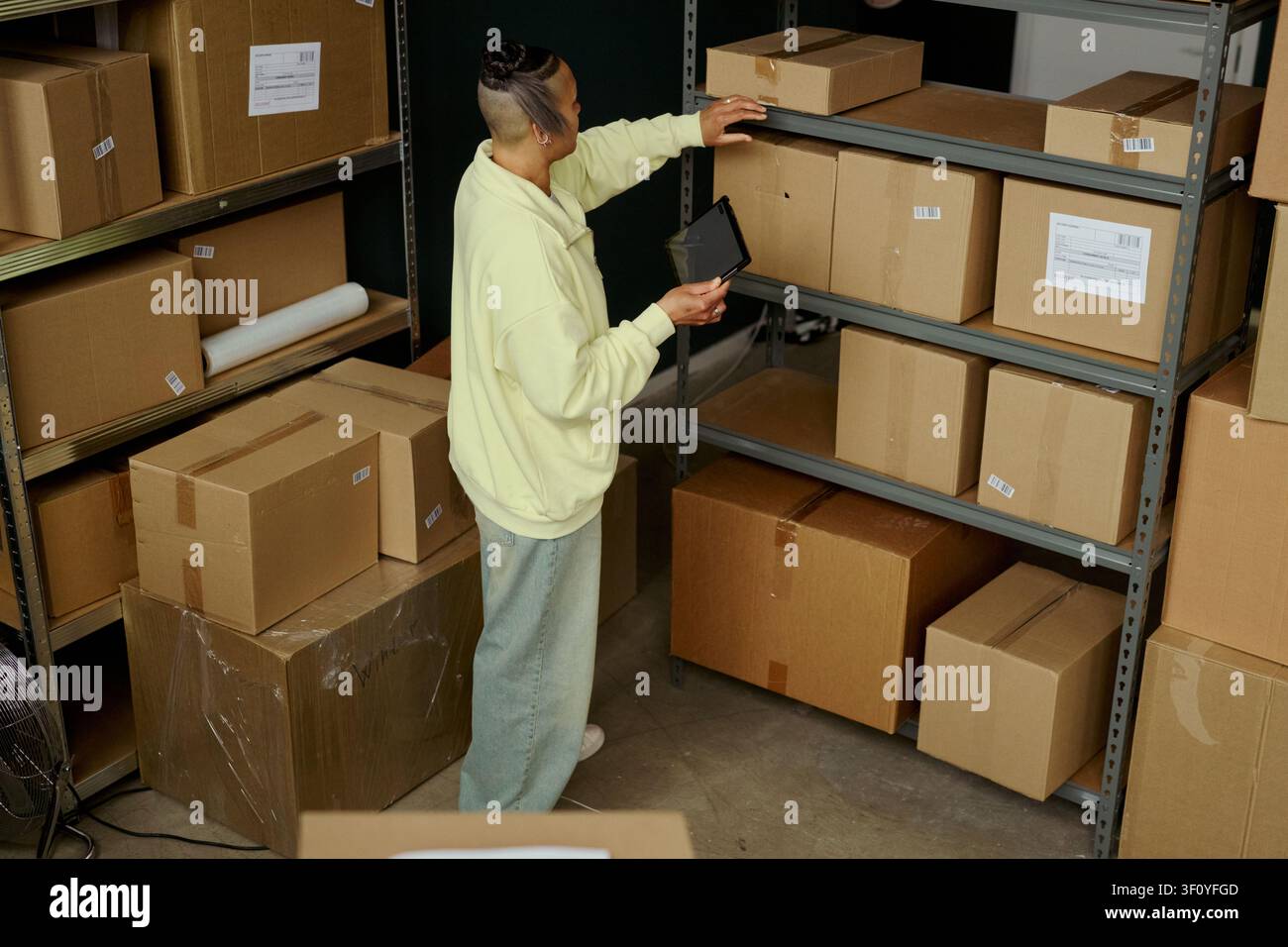 Hohe Blickwinkelansicht einer jungen erwachsenen biracialen Frau, die den Bestand auf einem digitalen Tablet überprüft, während sie nach einem Paket auf einem Lagerregal greift. Ideal für E-Commerce-Logistik, Lagerkontrolle, Erfüllungsmarketing Stockfoto