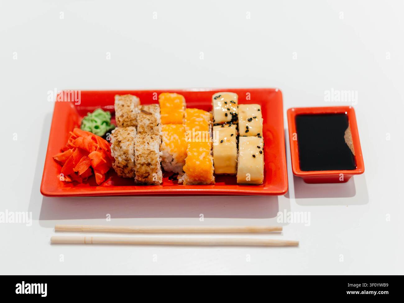 Frische verschiedene Sushi-Rollen, serviert auf einem lebhaften roten Teller mit Sojasauce, Wasabi, Ingwer und Stäbchen vor einem sauberen weißen Hintergrund. Traditionelle japanische Küche, farbenfroher Meeresfrüchte-Vorspeise, gesundes asiatisches Lebensmittelkonzept mit Platz für Text. Stockfoto