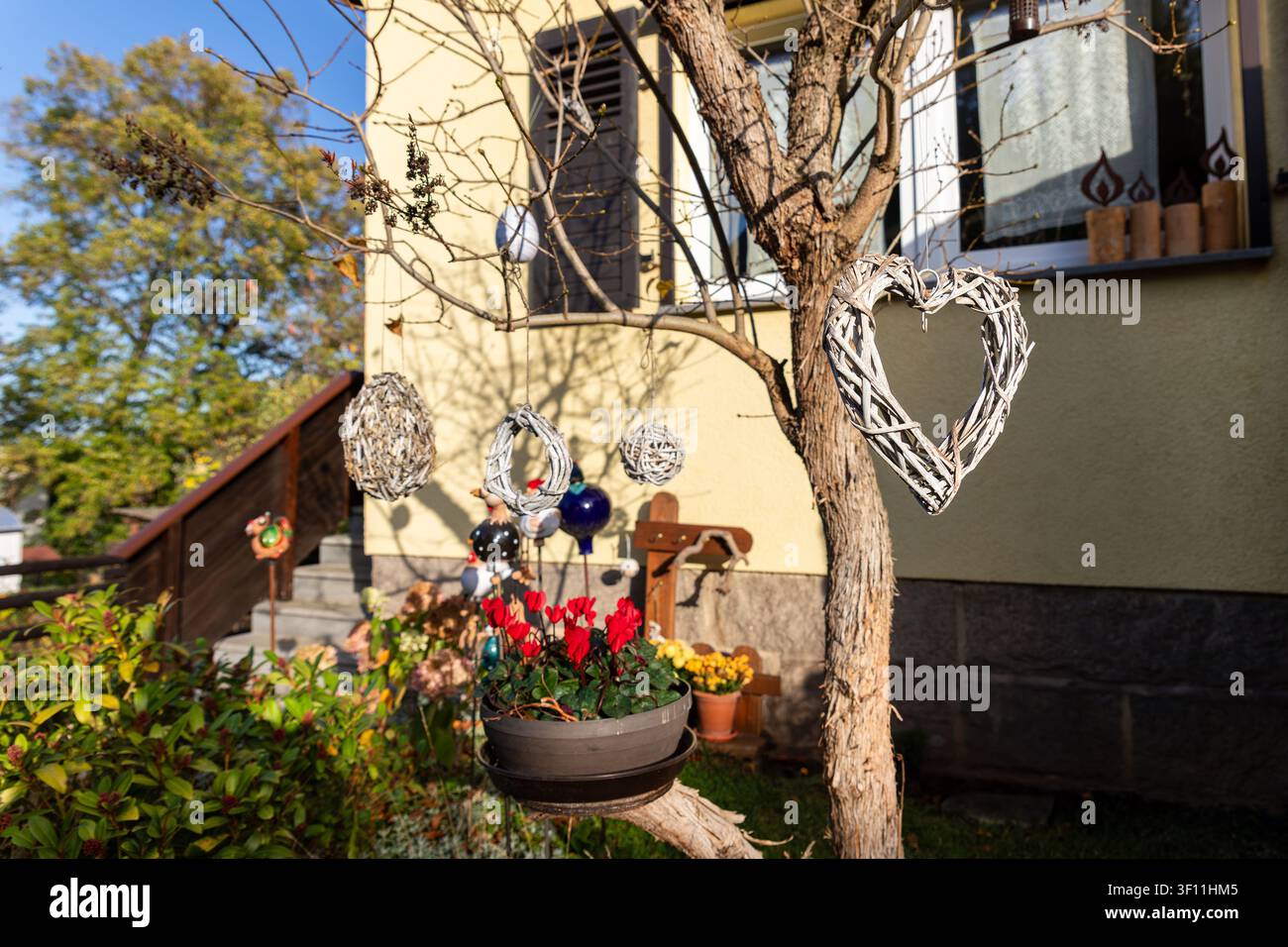 Rustikales Weidenherz hängt von Baumästen in einem sonnigen Hof neben bunten Blumen und einer gemütlichen Hausfassade. Handgemachte Ornamente, weicher Bokeh und warm Stockfoto