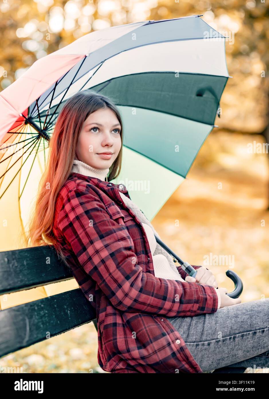 Eine junge Frau sitzt mit einem Regenschirm auf einer Parkbank. Sie wirkt entspannt, während die Blätter im Hintergrund die Farbe wechseln. Stockfoto