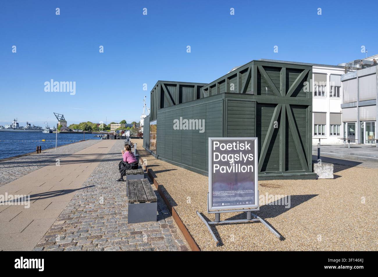 Eine Frau sitzt auf einer Holzbank am gepflasterten Kai der Galionsvej direkt neben den dunkelgrünen, gerüstartigen Holzkonstruktionen der vorübergehenden Architektur Stockfoto