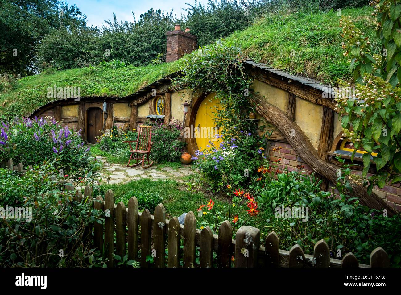 Ein malerischer Blick auf ein traditionelles Hobbit-Haus mit einer leuchtend gelben runden Tür, eingebettet in einen üppigen grünen Hügelgarten am Hobbiton-Filmset. Stockfoto