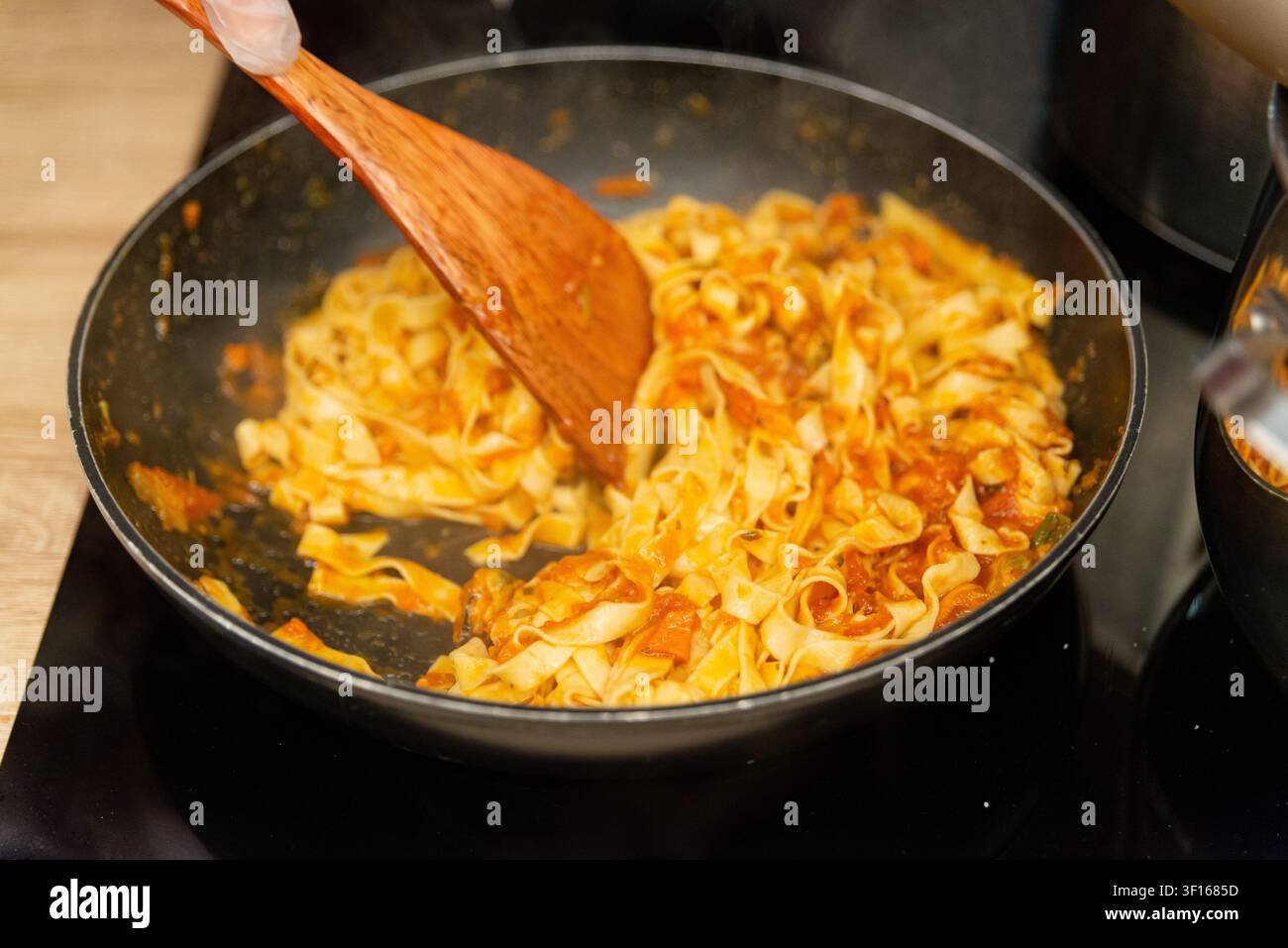 Nahaufnahme von dampfender Fettuccine-Pasta und Tomatensoße, die in einer schwarzen Pfanne mit einem hölzernen Spatel umgerührt wird. Stockfoto