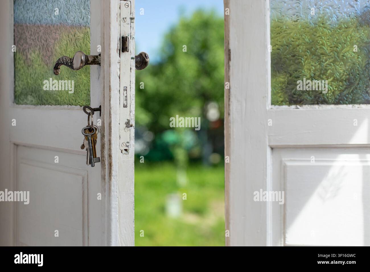 Alte offene weiße Holztür mit Milchglaspanelen und einem Schlüsselbund im Schloss, die zu einem hellen, unscharfen Sommergarten führt. Stockfoto
