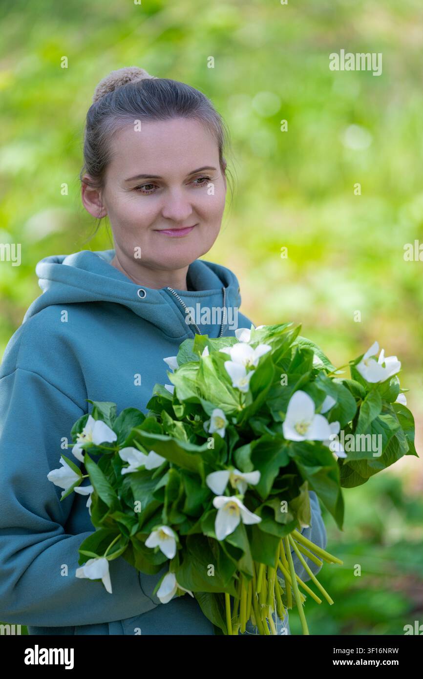 Weibliche Person in einem blaugrünen Hoodie, die frische Trilliumblüten mit üppigen grünen Blättern hält. Sanfte natürliche Beleuchtung, geringe Tiefenschärfe, ruhige Stimmung im Freien. Stockfoto
