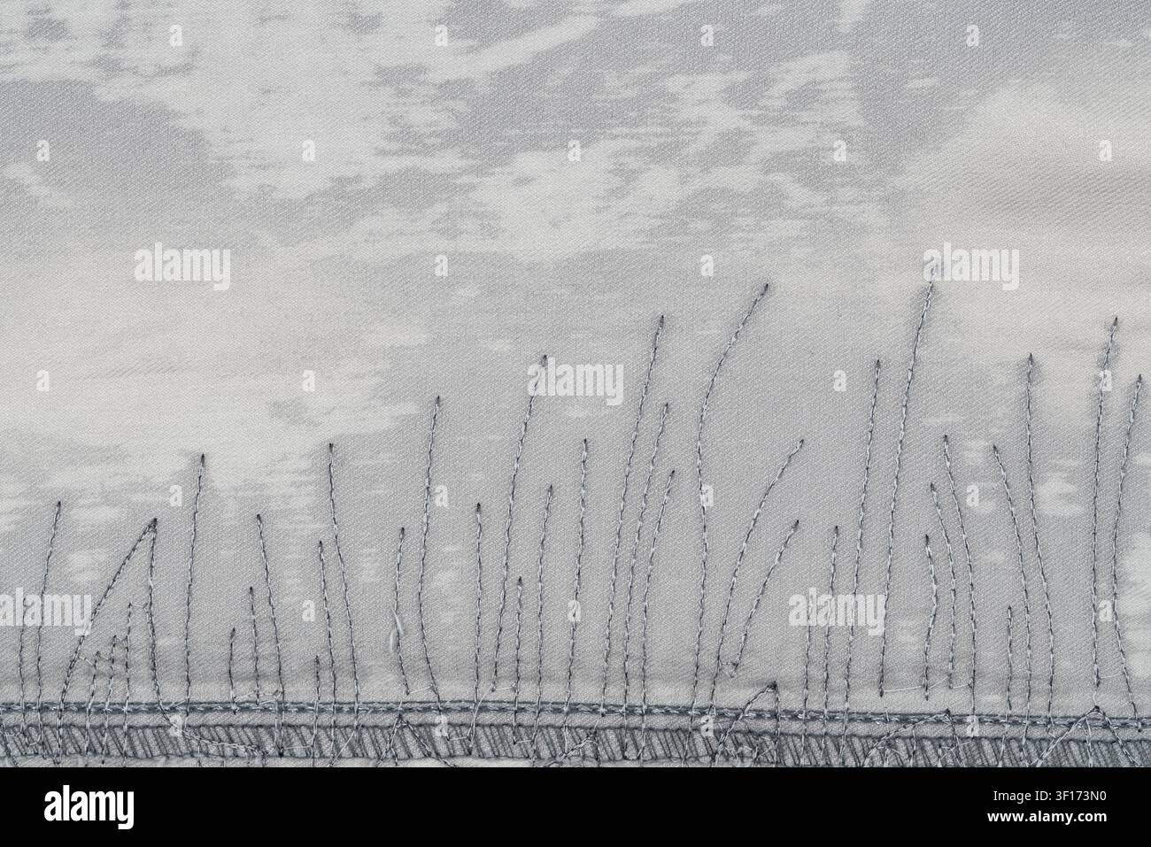 Nahaufnahme eines handgenähten Grasdesigns auf hellgrauem Stoff. Weiche Streiflichter betonen die Fadenstruktur, subtiler, wolkenartiger Hintergrund. Minimalist s Stockfoto